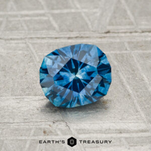 A 1.02-carat heated Montana sapphire gemstone is displayed on a textured gray surface with the "Earth's Treasury" logo and text at the bottom.