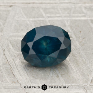 A 1.60-carat heated Montana sapphire, faceted in an oval cut with a deep blue hue, displayed on a textured gray surface above the text "EARTH'S TREASURY.