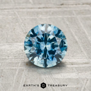A 1.16-carat heated Montana sapphire, round brilliant-cut and blue, is displayed on a textured gray surface with “Earth’s Treasury” at the bottom.