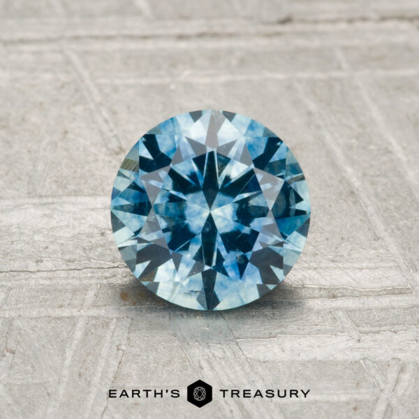 1.16-Carat Montana Sapphire (Heated) A 1.16-carat heated Montana sapphire, round brilliant-cut and blue, is displayed on a textured gray surface with “Earth’s Treasury” at the bottom.