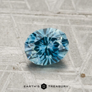 A 1.10-carat heated Montana sapphire with a faceted blue hue rests on a textured gray surface, with the text "EARTH'S TREASURY" displayed below the stone.