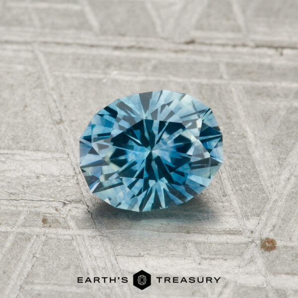 1.10-Carat Montana Sapphire (Heated) A 1.10-carat heated Montana sapphire with a faceted blue hue rests on a textured gray surface, with the text "EARTH'S TREASURY" displayed below the stone.