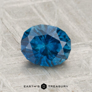 A 1.55-carat heated Montana sapphire is displayed on a textured gray surface with the text "EARTH'S TREASURY" below.