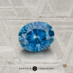 A 1.10-carat Montana sapphire (heated) with a faceted blue appearance rests on a textured metallic surface, featuring the text "Earth's Treasury" at the bottom.