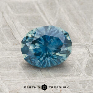 A faceted oval 1.47-carat heated Montana sapphire in blue rests on a textured gray surface, with the text "EARTH'S TREASURY" below.