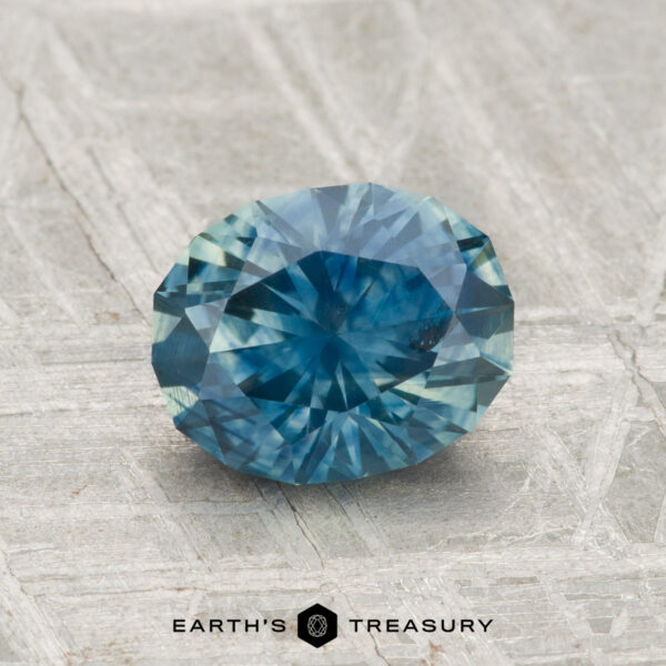 1.47-Carat Montana Sapphire (Heated) A faceted oval 1.47-carat heated Montana sapphire in blue rests on a textured gray surface, with the text "EARTH'S TREASURY" below.