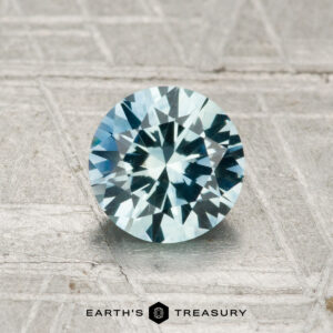 A 1.16-carat heated Montana sapphire with blue and green hues sits on a textured metallic surface, "Earth's Treasury" displayed below.