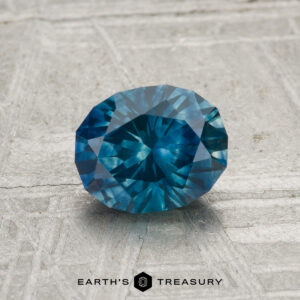 A 1.25-carat heated Montana sapphire, round and faceted, rests on a textured gray surface with "EARTH'S TREASURY" written at the bottom of the image.