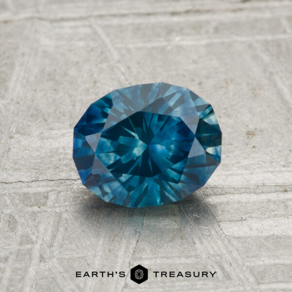 1.25-Carat Montana Sapphire (Heated) A 1.25-carat heated Montana sapphire, round and faceted, rests on a textured gray surface with "EARTH'S TREASURY" written at the bottom of the image.