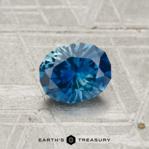 A 1.10-carat heated Montana sapphire with an oval cut is shown on a metallic, patterned surface featuring the "Earth's Treasury" logo at the bottom.