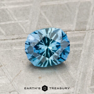 A 0.97-carat, cushion-cut blue Montana sapphire (heated) with intricate facets rests on a textured metallic surface. "EARTH'S TREASURY" appears at the bottom of the image.