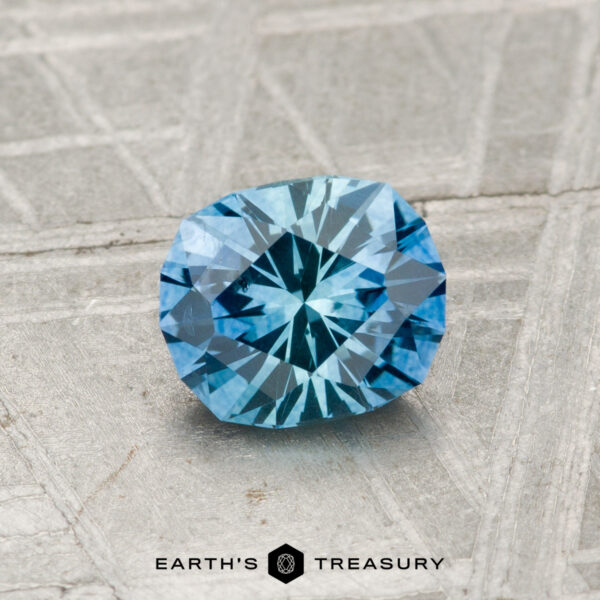 0.97-Carat Montana Sapphire (Heated) A 0.97-carat, cushion-cut blue Montana sapphire (heated) with intricate facets rests on a textured metallic surface. "EARTH'S TREASURY" appears at the bottom of the image.