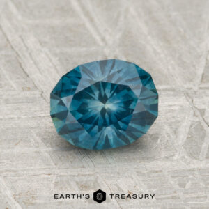 A 1.32-carat heated Montana sapphire, round and faceted with a teal-blue color, rests on a light gray textured surface; “EARTH'S TREASURY” appears at the bottom.