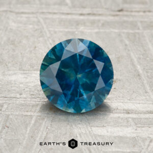 A 1.16-carat round-cut heated Montana sapphire is displayed on a textured, light surface with the "Earth's Treasury" logo and text beneath the gemstone.