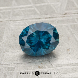 A 1.24-carat heated Montana sapphire, faceted in an oval cut, displayed on a textured gray surface with the Earth's Treasury logo at the bottom.