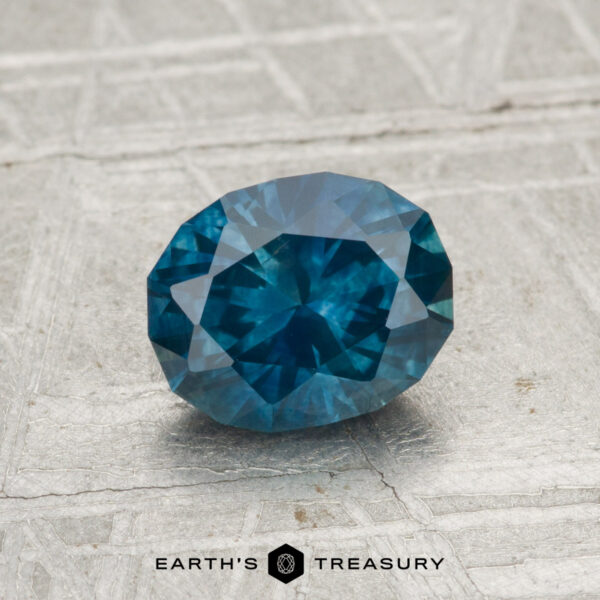 1.24-Carat Montana Sapphire (Heated) A 1.24-carat heated Montana sapphire, faceted in an oval cut, displayed on a textured gray surface with the Earth's Treasury logo at the bottom.