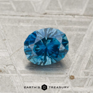 A 1.09-carat heated Montana sapphire, cut in a round brilliant style, rests on a textured gray surface with "EARTH'S TREASURY" displayed at the bottom.