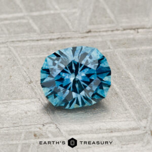 A 0.96-carat heated Montana sapphire rests on a textured metallic surface with "EARTH'S TREASURY" and a hexagon logo displayed below.
