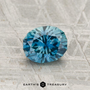 A 1.20-carat heated Montana sapphire, round and blue with facets, is shown on a textured gray surface featuring the "Earth's Treasury" logo at the bottom.