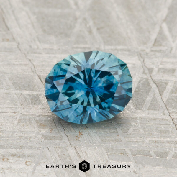 1.20-Carat Montana Sapphire (Heated) A 1.20-carat heated Montana sapphire, round and blue with facets, is shown on a textured gray surface featuring the "Earth's Treasury" logo at the bottom.