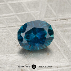 A 1.23-carat heated Montana sapphire with an oval cut rests on a textured metallic surface, "EARTH'S TREASURY" displayed at the bottom.