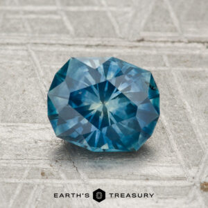 A faceted blue 1.34-carat heated Montana sapphire rests on a textured surface, with "Earth's Treasury" displayed at the bottom of the image.