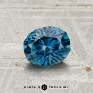 A 1.23-carat heated Montana sapphire, faceted in an oval shape and blue in color, is displayed on a textured gray surface with the "EARTH'S TREASURY" text and logo at the bottom.