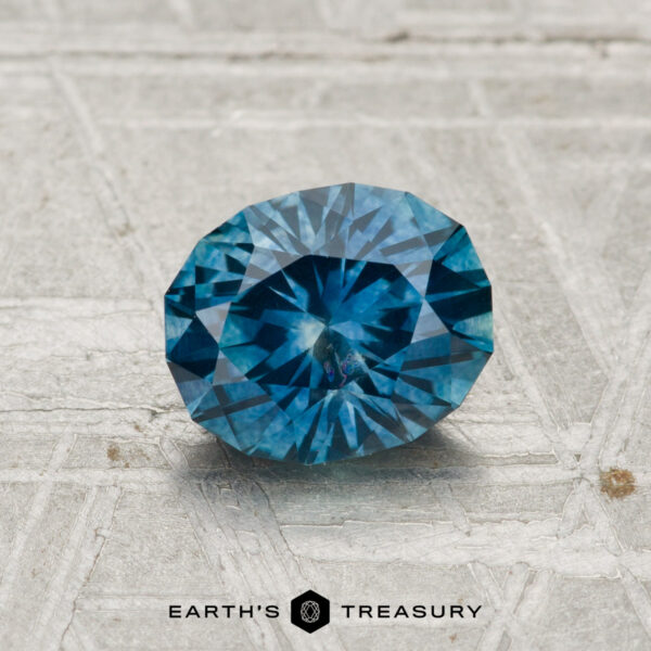 1.23-Carat Montana Sapphire (Heated) A 1.23-carat heated Montana sapphire, faceted in an oval shape and blue in color, is displayed on a textured gray surface with the "EARTH'S TREASURY" text and logo at the bottom.
