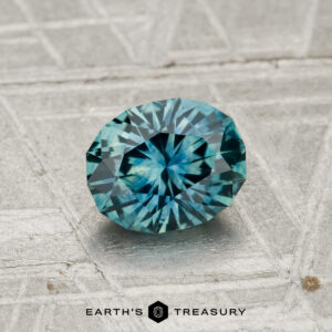 A 1.07-carat heated Montana sapphire shines in blue-green tones on a textured gray surface, with "EARTH'S TREASURY" displayed at the bottom.