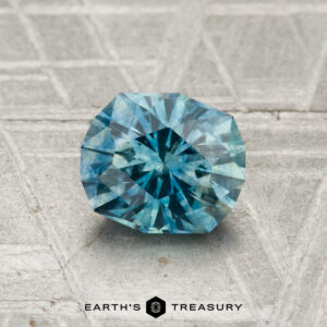 A 1.23-carat heated Montana sapphire, round and blue-green with faceted cuts, rests on a textured metallic surface. "EARTH'S TREASURY" is displayed at the bottom.