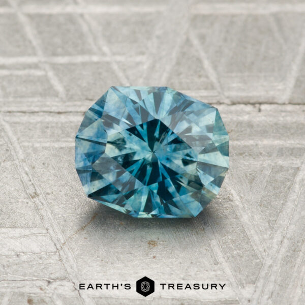 1.23-Carat Montana Sapphire (Heated) A 1.23-carat heated Montana sapphire, round and blue-green with faceted cuts, rests on a textured metallic surface. "EARTH'S TREASURY" is displayed at the bottom.