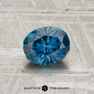 A 1.06-carat heated Montana Sapphire with blue facets is displayed on a textured gray surface, featuring the "EARTH'S TREASURY" name and logo at the bottom of the image.