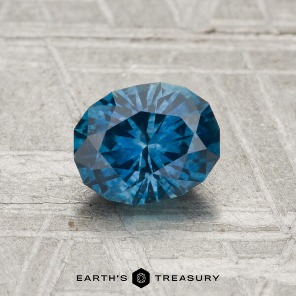 1.06-Carat Montana Sapphire (Heated) A 1.06-carat heated Montana Sapphire with blue facets is displayed on a textured gray surface, featuring the "EARTH'S TREASURY" name and logo at the bottom of the image.
