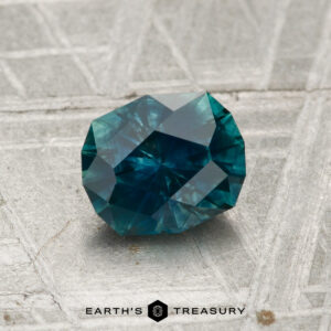 A 1.19-carat heated Montana sapphire, faceted in blue-green hues, rests on a textured metallic surface with the "Earth's Treasury" logo and text below.