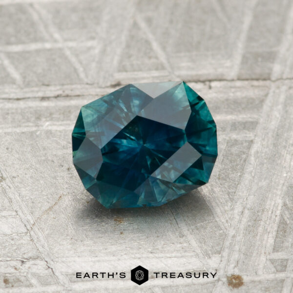 1.19-Carat Montana Sapphire (Heated) A 1.19-carat heated Montana sapphire, faceted in blue-green hues, rests on a textured metallic surface with the "Earth's Treasury" logo and text below.