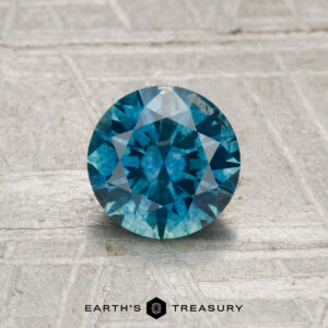 A 1.11-carat heated Montana sapphire, round brilliant-cut and blue, displayed on a textured gray surface with the "Earth's Treasury" logo and text at the bottom.