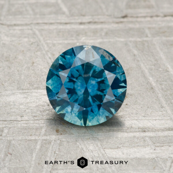 1.11-Carat Montana Sapphire (Heated) A 1.11-carat heated Montana sapphire, round brilliant-cut and blue, displayed on a textured gray surface with the "Earth's Treasury" logo and text at the bottom.