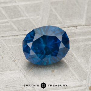A 1.23-carat heated Montana sapphire, oval-shaped and blue, rests on a textured metallic surface with the "Earth's Treasury" logo at the bottom.