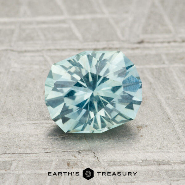 1.37-Carat Montana Sapphire A 1.37-carat Montana Sapphire, faceted in a light aqua blue hue, is displayed on a textured gray background with the text “Earth’s Treasury” at the bottom.