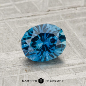 A faceted, round brilliant-cut 1.21-carat heated Montana sapphire is displayed on a textured gray surface with the text "EARTH'S TREASURY" below.