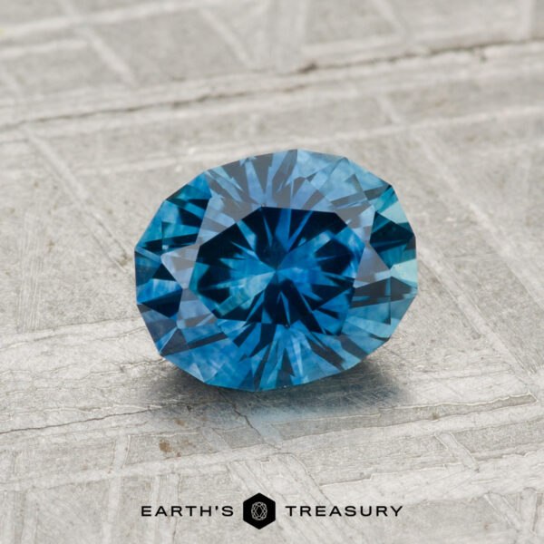 1.21-Carat Montana Sapphire (Heated) A faceted, round brilliant-cut 1.21-carat heated Montana sapphire is displayed on a textured gray surface with the text "EARTH'S TREASURY" below.