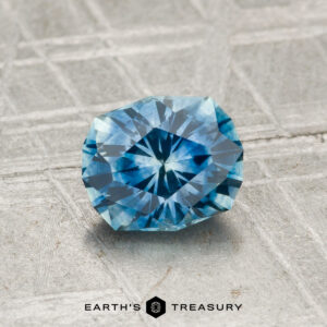 A faceted blue 1.18-carat heated Montana sapphire with geometric patterns rests on a textured gray surface; the "Earth's Treasury" logo is visible at the bottom.