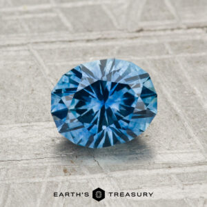 A 1.21-carat heated Montana sapphire with a round brilliant cut is showcased on a textured gray surface, with "Earth's Treasury" displayed at the bottom.
