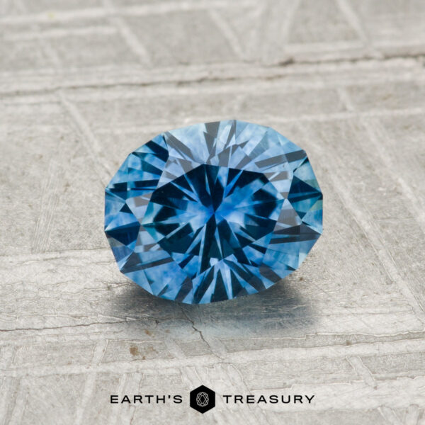 1.21-Carat Montana Sapphire (Heated) A 1.21-carat heated Montana sapphire with a round brilliant cut is showcased on a textured gray surface, with "Earth's Treasury" displayed at the bottom.