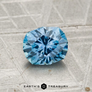 A 1.10-carat heated Montana sapphire, precision-cut in a round brilliant style, shown on a textured gray surface with the "Earth's Treasury" logo at the bottom.