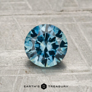 A round, faceted 1.10-carat heated Montana sapphire with a blue-green hue rests on a textured, light-colored surface, with "EARTH'S TREASURY" displayed at the bottom.
