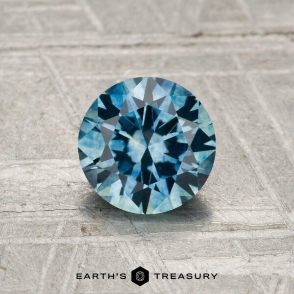 1.10-Carat Montana Sapphire (Heated) A round, faceted 1.10-carat heated Montana sapphire with a blue-green hue rests on a textured, light-colored surface, with "EARTH'S TREASURY" displayed at the bottom.
