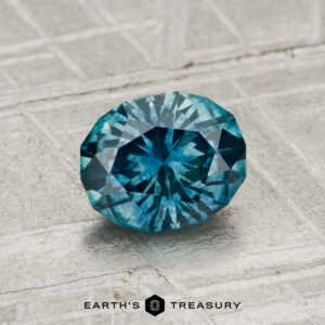 A 1.21-carat heated Montana sapphire with a round brilliant cut is displayed on a textured gray surface, featuring the text "EARTH'S TREASURY" at the bottom.