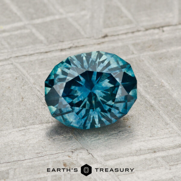 1.21-Carat Montana Sapphire (Heated) A 1.21-carat heated Montana sapphire with a round brilliant cut is displayed on a textured gray surface, featuring the text "EARTH'S TREASURY" at the bottom.