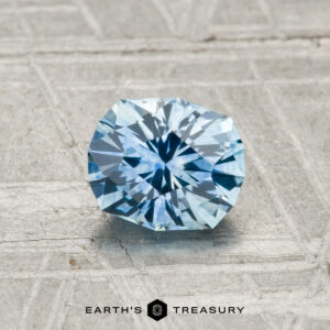 A 0.96-carat heated Montana Sapphire, round and faceted in light blue, is displayed on a gray textured surface with "EARTH'S TREASURY" text at the bottom.